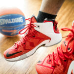 Gros plan sur des chaussures de basketball rouges portées sur un terrain en parquet, avec un ballon de basket en arrière-plan.
