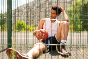 Jeune homme actif et décontracté assis près de la clôture d’un terrain de basket, tenant un ballon pendant une courte pause.