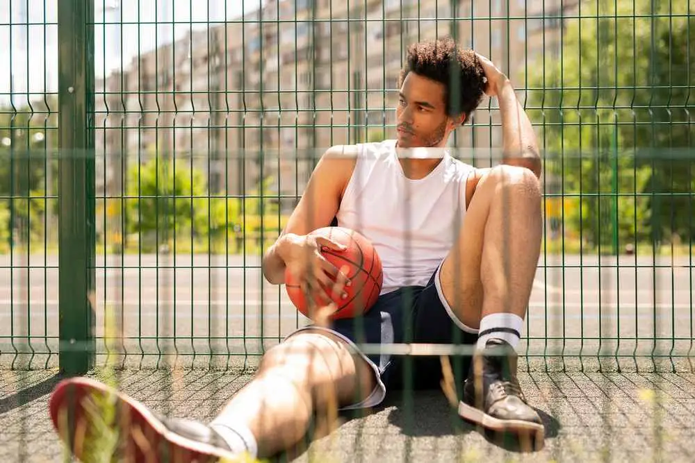 Jeune joueur de basketball assis au bord d'un terrain extérieur avec son ballon pendant une pause après l'entraînement.